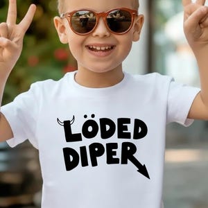 May include: A white t-shirt with the text "LÖDED DIPER" in black, with a devil horn above the "L" and an arrow pointing down from the "R". The person in the image is wearing brown sunglasses and making a peace sign.