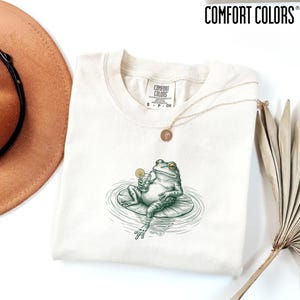 May include: A cream-colored t-shirt with a Comfort Colors brand label at the neck. The shirt features a graphic of a green frog sitting on a lily pad, holding a drink with a lemon slice. A gold-toned necklace rests on the fabric of the shirt.