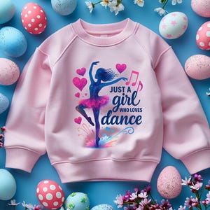 May include: A light pink sweatshirt featuring a ballerina graphic with the text "Just a girl who loves dance." The design includes hearts and musical notes. The background includes decorated Easter eggs and flowers.