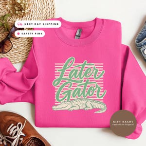 May include: A bright pink crewneck sweatshirt featuring a graphic of a light-colored alligator positioned below the text "Later Gator" in a cursive green font, set against a background of horizontal light pink stripes on the front of the garment.