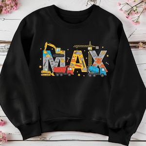 May include: Black sweatshirt featuring the name "MAX" in colorful construction-themed lettering. The letters are designed with images of construction vehicles, tools, and building materials. The design includes a yellow excavator, a red dump truck, and a blue cement mixer.