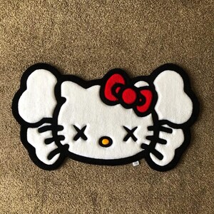 May include: White rug shaped like a cartoon cat's face with a red bow, black outline, and black cross-shaped eyes. The rug has a fluffy texture and is placed on a brown carpet.