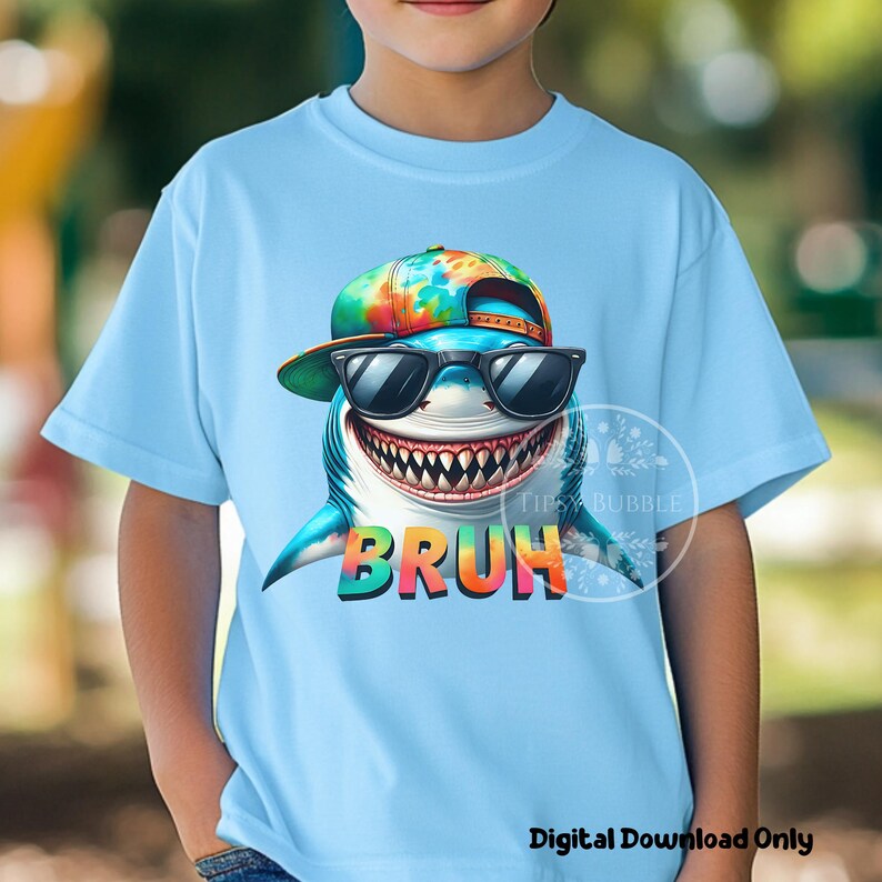 May include: A light blue t-shirt with a colorful graphic of a shark wearing sunglasses and a tie-dye hat. The shark's mouth is open and the text "BRUH" is printed below the shark.