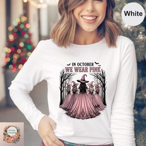 May include: A white long-sleeve shirt featuring a graphic of five witches in pink dresses and hats standing in a forest. The text above the illustration reads IN OCTOBER WE WEAR PINK, accompanied by small black bat silhouettes on either side.
