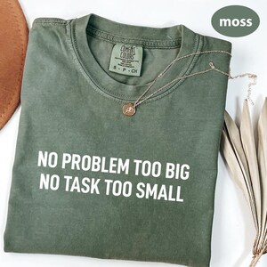 May include: A moss green t-shirt with the text "NO PROBLEM TOO BIG NO TASK TOO SMALL" in white. A gold necklace with a pendant rests on the shirt. The shirt has a Comfort Colors tag.