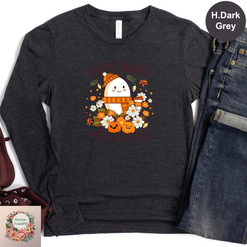 a gray long-sleeved t-shirt with a graphic design of a ghost surrounded by autumn-themed elements, such as pumpkins, leaves, and flowers. The t-shirt is displayed alongside a pair of jeans and a pair of shoes, suggesting a casual outfit.