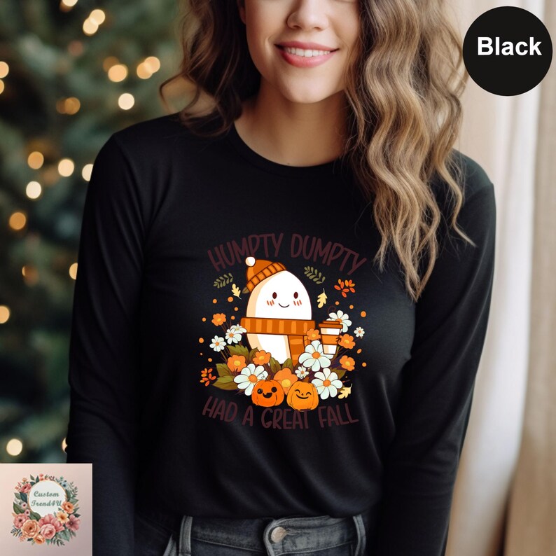 A woman wearing a black long-sleeved shirt with a graphic design featuring a ghost surrounded by autumn-themed elements, including pumpkins, flowers, and leaves.