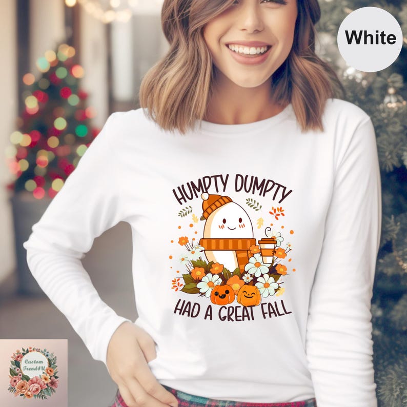 A woman wearing a white long-sleeved shirt with a graphic design featuring a ghost, pumpkins, and text that says "Humpty Dumpty had a great fall".
