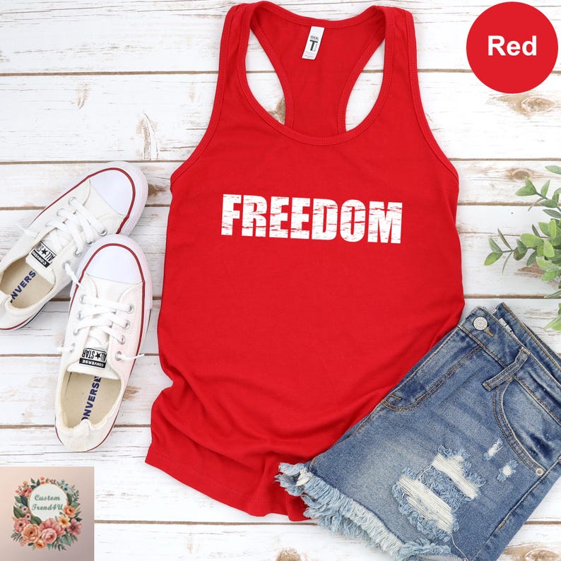 a red tank top with the word "FREEDOM" printed on it, a pair of white sneakers, and a pair of blue denim shorts, all laid out on a white wooden surface.