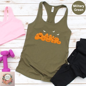 May include: Military green tank top with a pumpkin graphic. The sleeveless top features a row of orange pumpkins with smiling faces and small bats. A pink sports bra and weights are visible in the background.