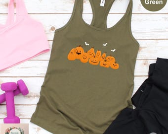 May include: Military green tank top with a pumpkin graphic. The sleeveless top features a row of orange pumpkins with smiling faces and small bats. A pink sports bra and weights are visible in the background.