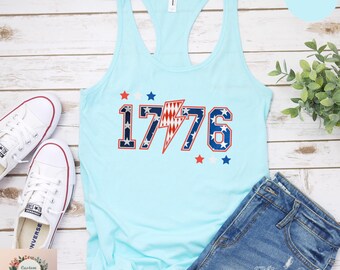 May include: Light blue tank top with the number "1776" in red, white, and blue, with a lightning bolt graphic. The tank top is paired with denim shorts and white sneakers. The design incorporates stars.