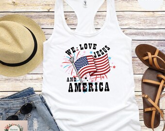 May include: White tank top with a patriotic design. The graphic features the American flag and the words "We Love Jesus and America" in black. The tank top is a racerback style.