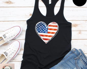 May include: Black racerback tank top with a heart-shaped American flag design. The heart features the stars and stripes of the US flag, outlined in white. The tank top is paired with denim shorts and white sneakers.