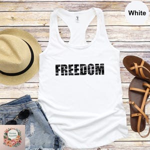 a white tank top with the word "FREEDOM" printed on it, along with a straw hat, denim shorts, and sandals arranged on a wooden surface.