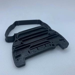 May include: A black, 3D-printed object with a unique shape, featuring a handle-like strap. The main body has a textured surface with geometric details. The design suggests a tool or accessory.