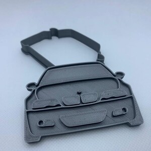 May include: A gray 3D-printed car-shaped cookie cutter. The cutter features the front of a car with headlights, a grill, and a handle at the top. The design is detailed and shows the car's shape.