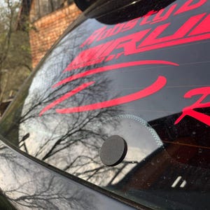 Puede incluir: Primer plano de una ventana de coche negra con calcomanías rojas. Las calcomanías incluyen la palabra "RACING" y otros textos. Un pequeño objeto redondo y negro está adherido al cristal. El fondo muestra árboles y un edificio.