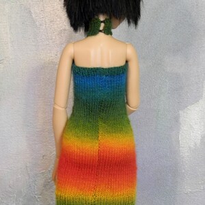 May include: A doll wearing a rainbow knit dress with a green, blue, yellow, orange, and red color gradient. The dress is sleeveless and has a straight neckline.
