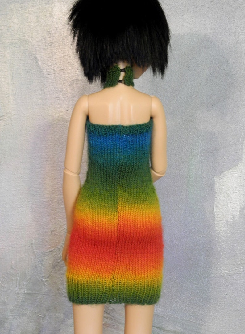 May include: A doll wearing a rainbow knit dress with a green, blue, yellow, orange, and red color gradient. The dress is sleeveless and has a straight neckline.
