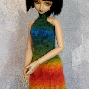May include: A doll wearing a rainbow knit dress with a high neck and a sleeveless design. The dress is a gradient of green, blue, yellow, orange, and red.