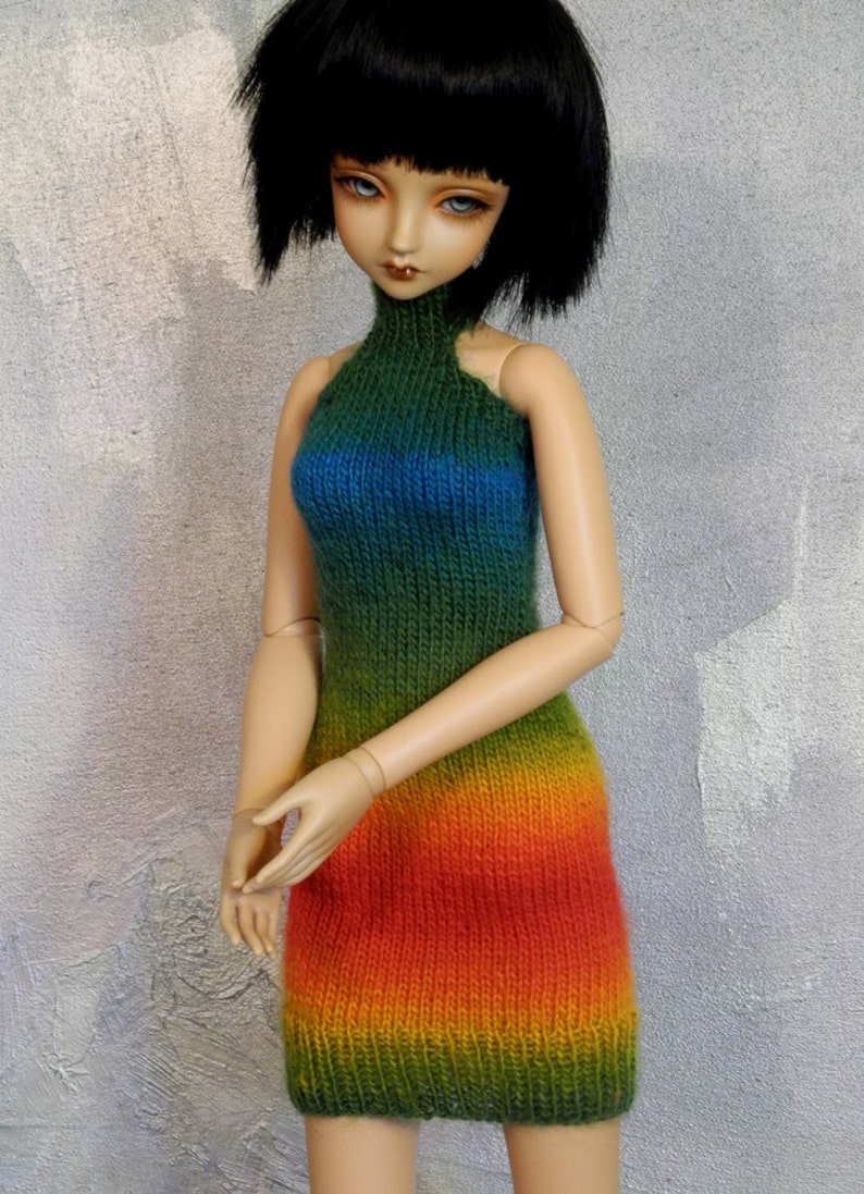 May include: A doll wearing a rainbow knit dress with a high neck and a sleeveless design. The dress is a gradient of green, blue, yellow, orange, and red.