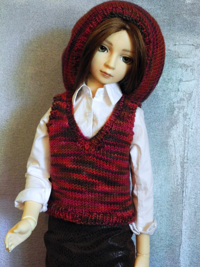 May include: A doll wearing a red and brown striped knitted waistcoat, a white button-down shirt, and a red knitted hat.