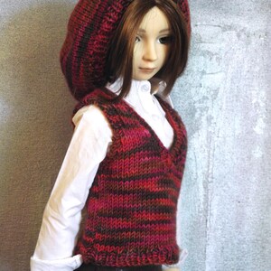 May include: A doll wearing a red and brown striped knitted waistcoat and a matching knitted hat. The doll has long brown hair and is wearing a white shirt.