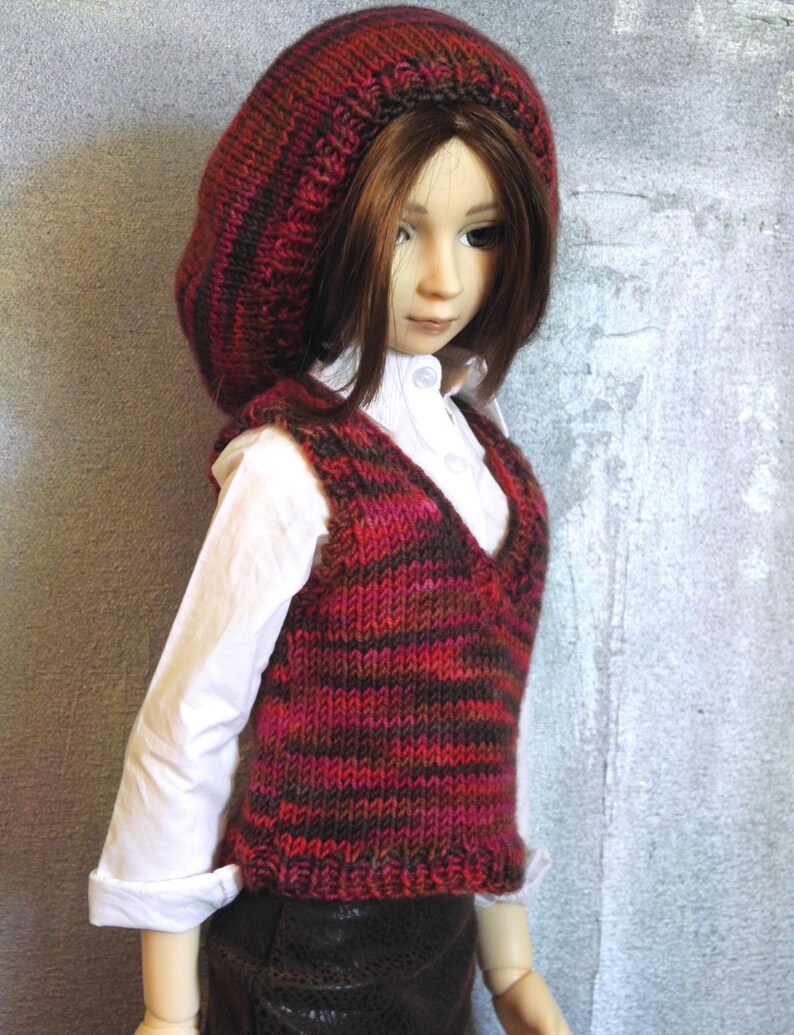 May include: A doll wearing a red and brown striped knitted waistcoat and a matching knitted hat. The doll has long brown hair and is wearing a white shirt.