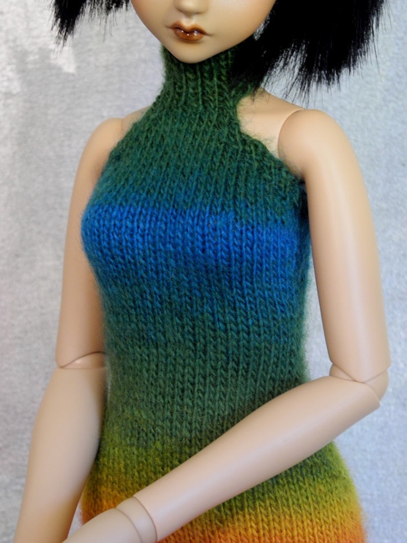 May include: A doll wearing a green, blue, and orange ombre knitted halter top.