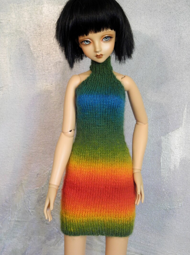 May include: A doll wearing a rainbow knit halter dress. The dress is green, blue, yellow, orange, and red.