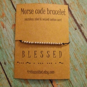 May include: A brown card with the text "Morse code bracelet" and "stainless steel & waxed cotton cord". A silver bracelet with the word "BLESSED" spelled out in Morse code is on the card.