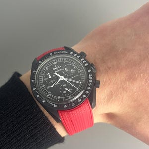 May include: A black and red chronograph wristwatch with a black rubber strap. The watch face has a tachymeter scale and the words "OMEGA Swatch" printed on it.