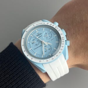 May include: A light blue wristwatch with a white strap. The watch face is white with black markings and the text "OMEGA Speedmaster Moon Watch" printed on it.