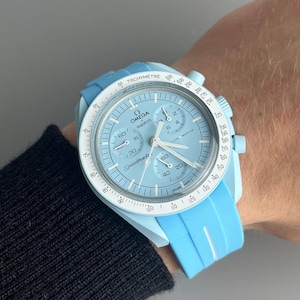Peut inclure: Une montre-bracelet bleu clair avec un cadran blanc et des aiguilles noires. La montre a une échelle tachymétrique autour du bord du cadran et les mots "Omega swatch" imprimés sur le cadran. La montre est sur un bracelet en silicone bleu clair.