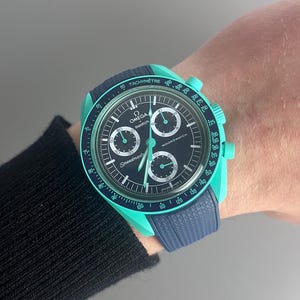 Puede incluir: Un reloj de pulsera cronógrafo Omega Speedmaster Moonwatch de color turquesa y negro con una correa de nailon azul. La esfera del reloj tiene una escala de taquímetro y las palabras "Speedmaster" y "Moonwatch" impresas en ella.
