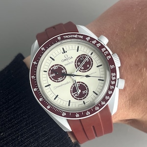 May include: A white and claret Omega Speedmaster watch with a claret strap. The watch face has black numerals and sub-dials. The watch has the text "TACHYMÈTRE" and "Speedmaster".