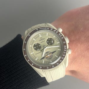 May include: A white and brown wristwatch with a chronograph face. The watch has a white strap with a textured pattern. The watch face has the words "Omega" and "Speedmaster" printed on it.