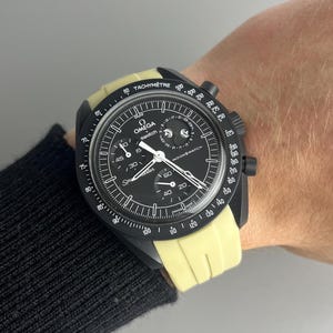 May include: A black and yellow Omega x Swatch MoonSwatch Speedmaster watch with a tachymeter scale on the bezel. The watch has a black face with white markings and a date window at the 3 o'clock position. The watch is on a person's wrist.