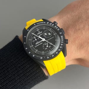 May include: A black and yellow wristwatch with a black face and a tachymeter scale. The watch face has the words "OMEGA" and "Speedmaster" printed on it. The watch is on a yellow silicone strap.
