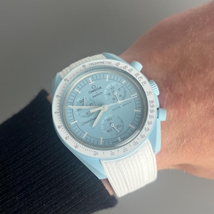 Peut inclure: Une montre-bracelet bleu clair avec un bracelet blanc. Le cadran de la montre comporte une échelle tachymétrique blanche, une aiguille des secondes blanche et les mots "OMEGA" et "Speedmaster" imprimés dessus.