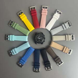 Op de afbeelding: Een assortiment van horlogebandjes in verschillende kleuren, waaronder rood, geel, blauw, groen en grijs. De bandjes zijn in een cirkel rond een zwart horloge gezicht gerangschikt.