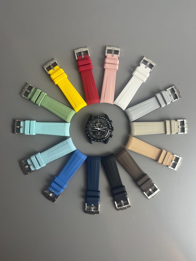 Op de afbeelding: Een assortiment van horlogebandjes in verschillende kleuren, waaronder rood, geel, blauw, groen en grijs. De bandjes zijn in een cirkel rond een zwart horloge gezicht gerangschikt.