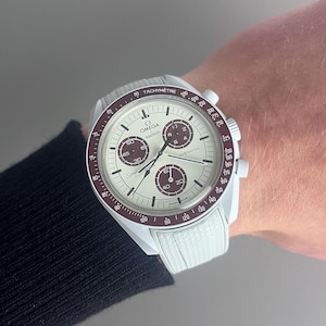 May include: A white and red chronograph wristwatch with a white nylon strap. The watch face has a white background with black markings and a red second hand. The watch face also has the words "Omega" and "Swatch" printed on it.