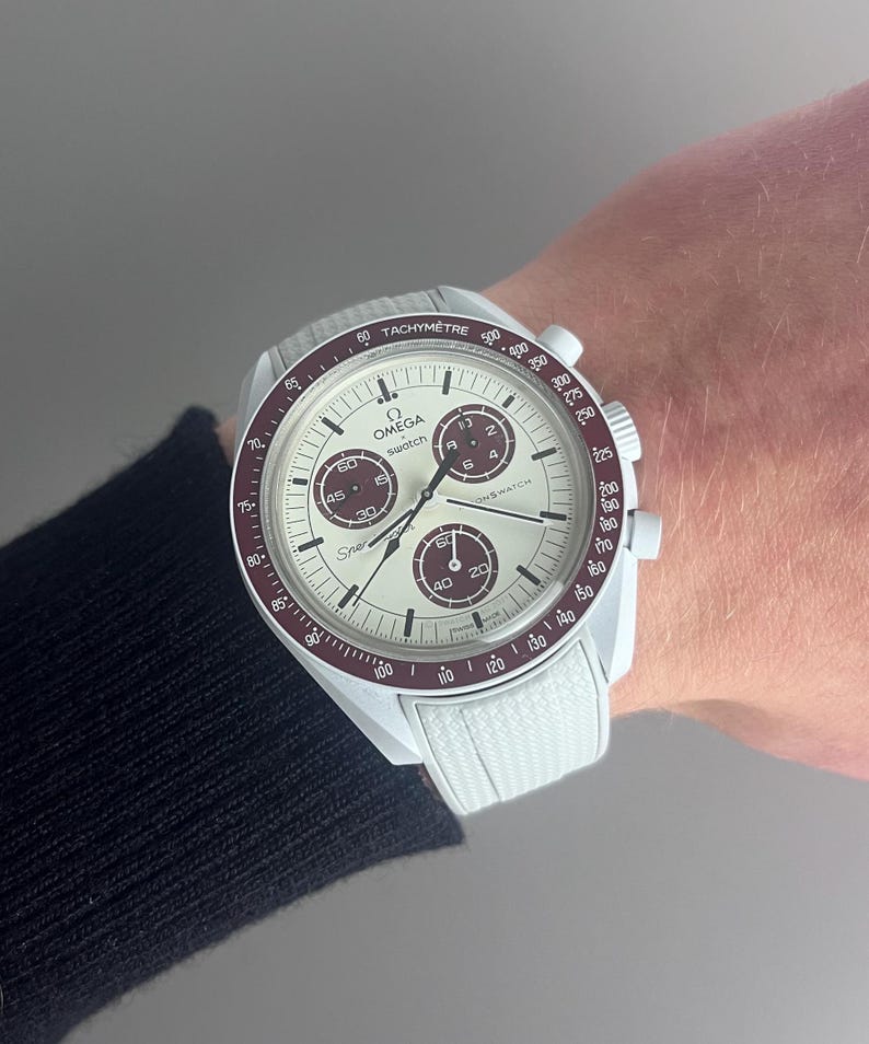 Op de afbeelding: Een wit en rood chronograaf polshorloge met een witte nylon band. De wijzerplaat van het horloge heeft een witte achtergrond met zwarte markeringen en een rode secondewijzer. Op de wijzerplaat van het horloge staan ook de woorden "Omega" en "Swatch" gedrukt.