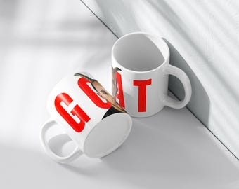 May include: Two white ceramic mugs with the word "GOAT" printed in red on them. One mug has a person's face on it, while the other mug has a plain white surface.