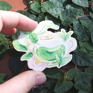 Puede incluir: Una ilustración de una tetera y una taza de té verdes con flores blancas y hojas verdes. La ilustración está sobre un fondo blanco.