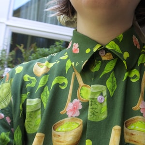 Puede incluir: Una camisa verde de botones con un patrón de cuencos de té matcha, batidores y flores. La camisa tiene cuello y cierre de botones.