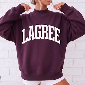 May include: A dark plum-colored long-sleeve sweatshirt featuring a mock neck collar and ribbed cuffs. The word LAGREE is printed in large, white, arched capital letters across the center of the chest. The garment has a relaxed, loose fit.