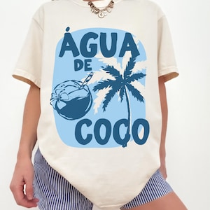 May include: Cream-colored t-shirt featuring a blue graphic with the words "AGUA DE COCO". The design includes a coconut drink and a palm tree silhouette, evoking a tropical theme. The shirt has short sleeves.